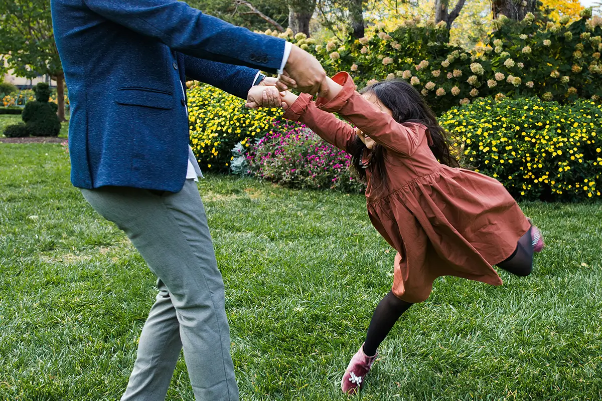 How Much Does DHS Pay for Child Care in Oklahoma? - Child Care Inc An adult spins a young girl by her hands in a grassy, flower-filled park.