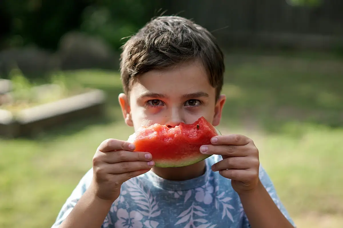 A young boy holding a piece of half-eaten watermelon above his mouth